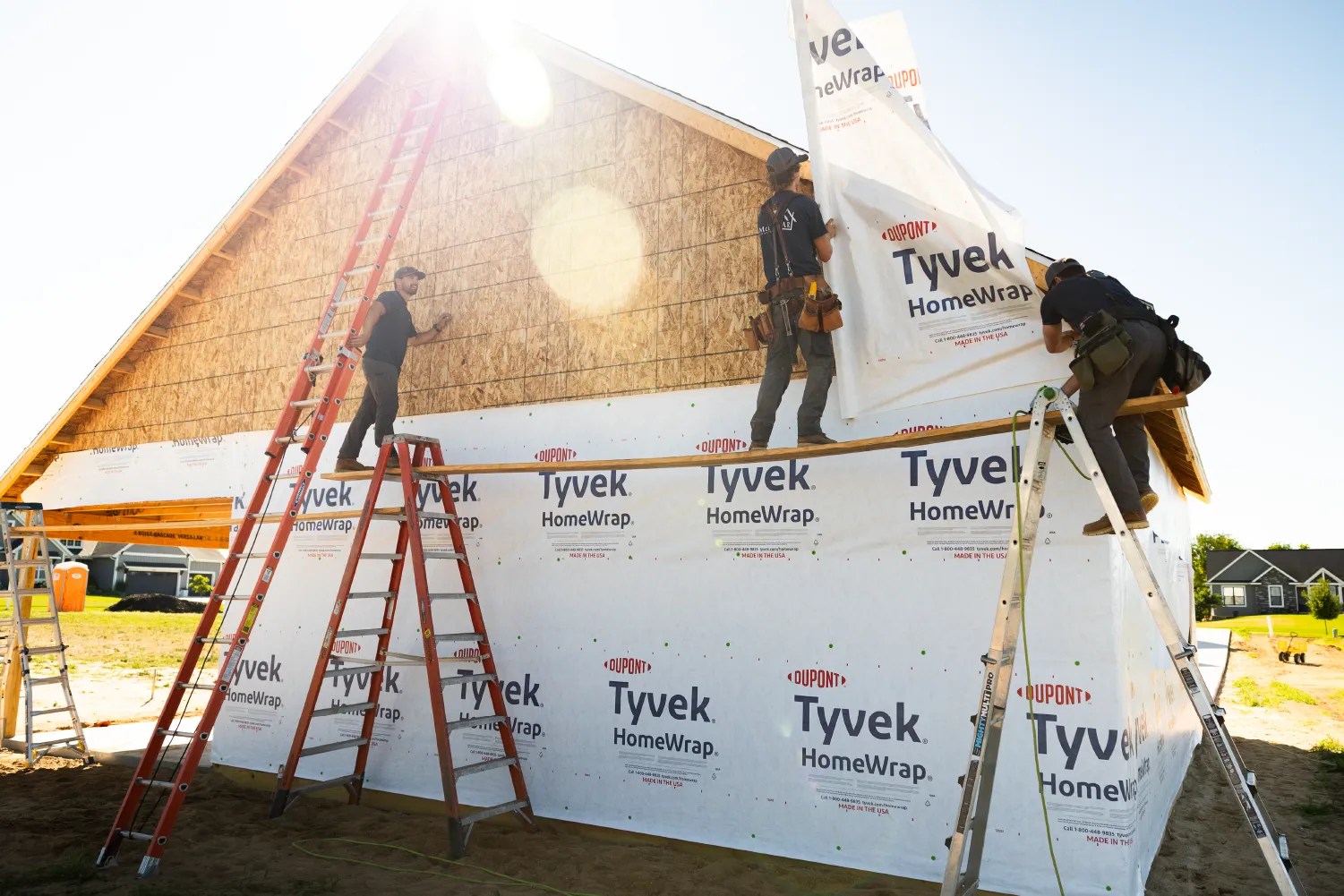 Three construction workers installing Tyvek HomeWrap on the exterior of a wooden framed building under clear sky.