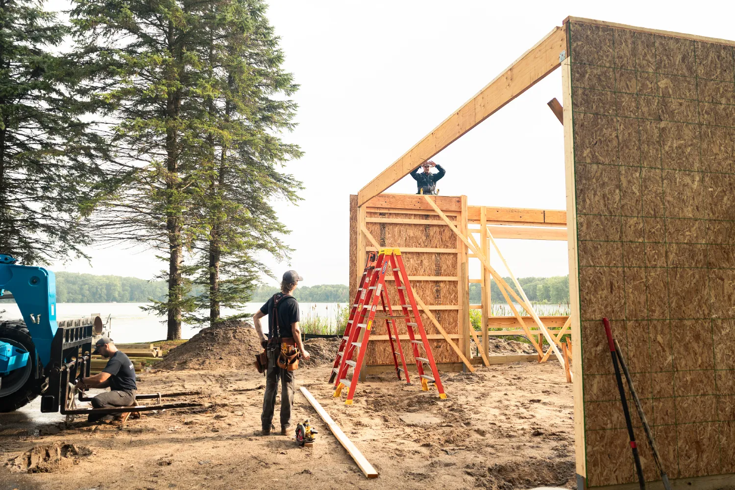 Three construction workers building a wooden framed structure near a lake with trees in the background.