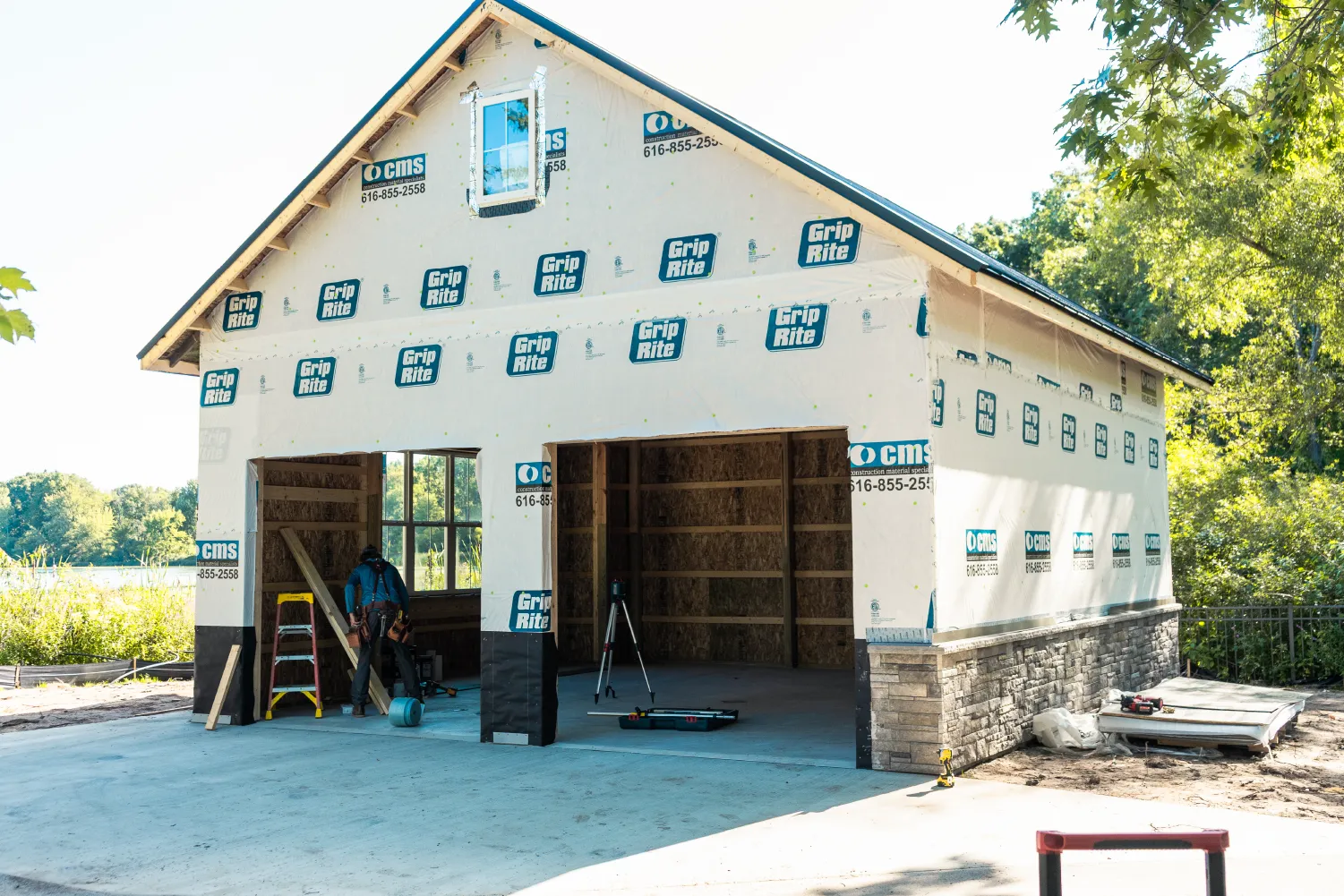 Partially constructed barn with weatherproof wrap and stone siding, a worker inside, and construction tools around.