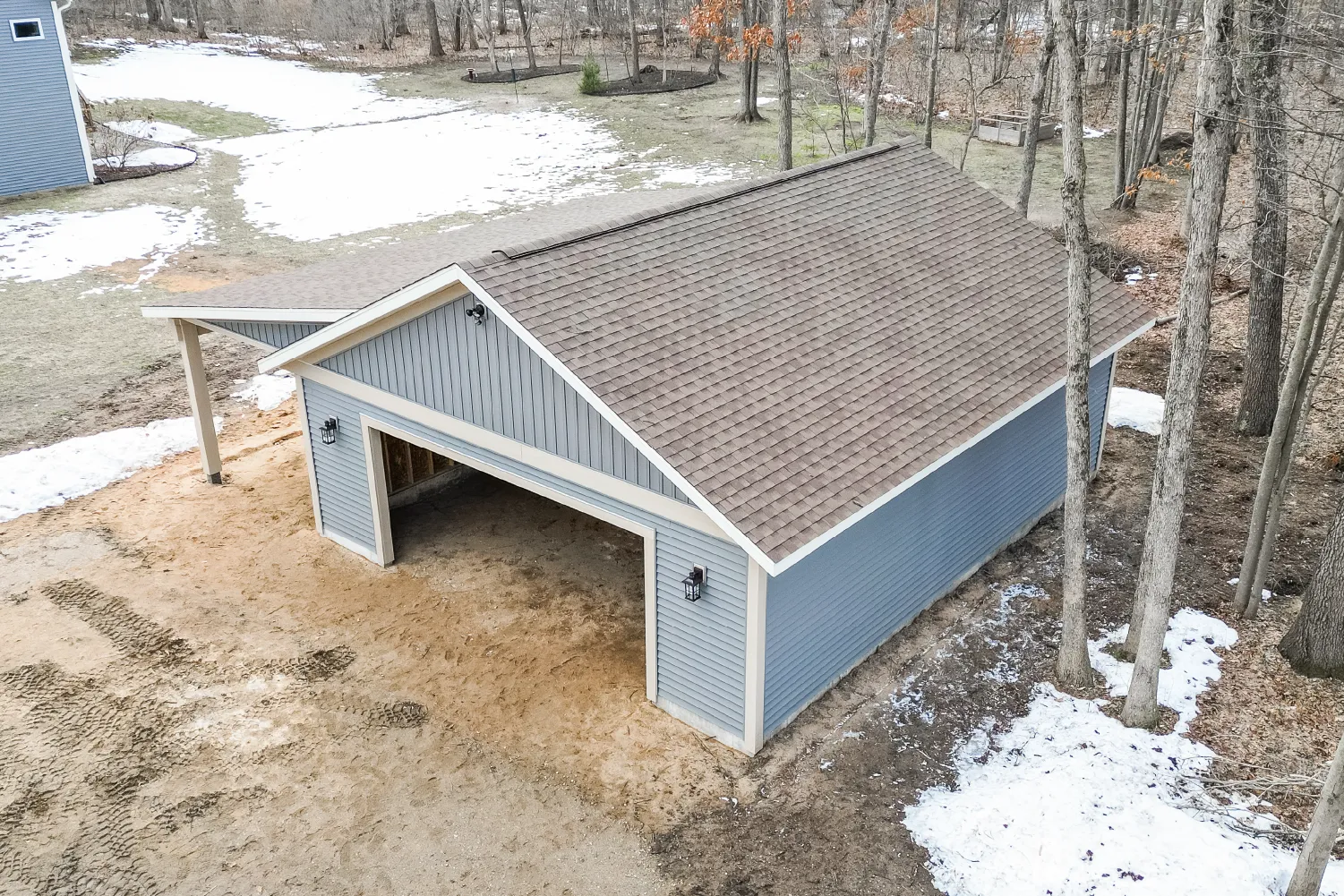 Newly constructed blue two-car garage with brown shingle roof in a lightly snowy yard surrounded by trees.