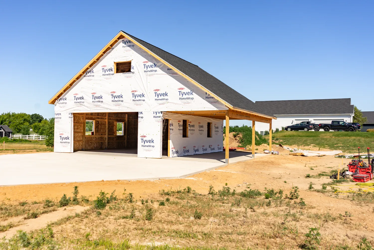 Framed barn under construction with Tyvek HomeWrap and a concrete driveway, set in a rural area with clear blue sky.