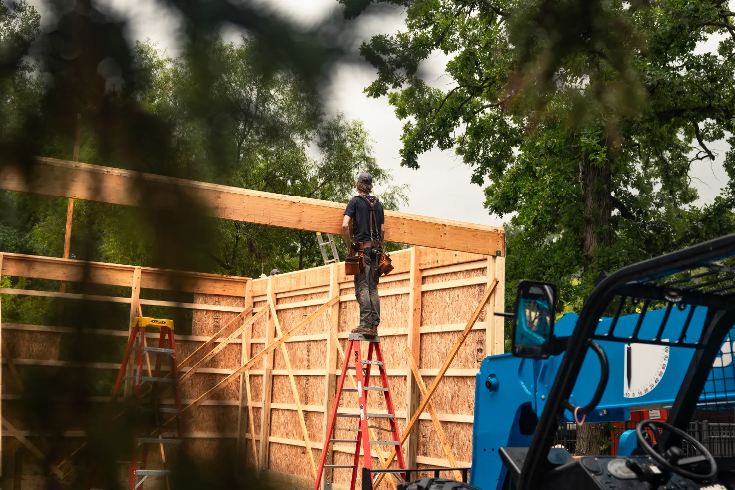 Construction worker standing on a red ladder installing a wooden beam on a partially built wooden structure outdoors with trees in background.