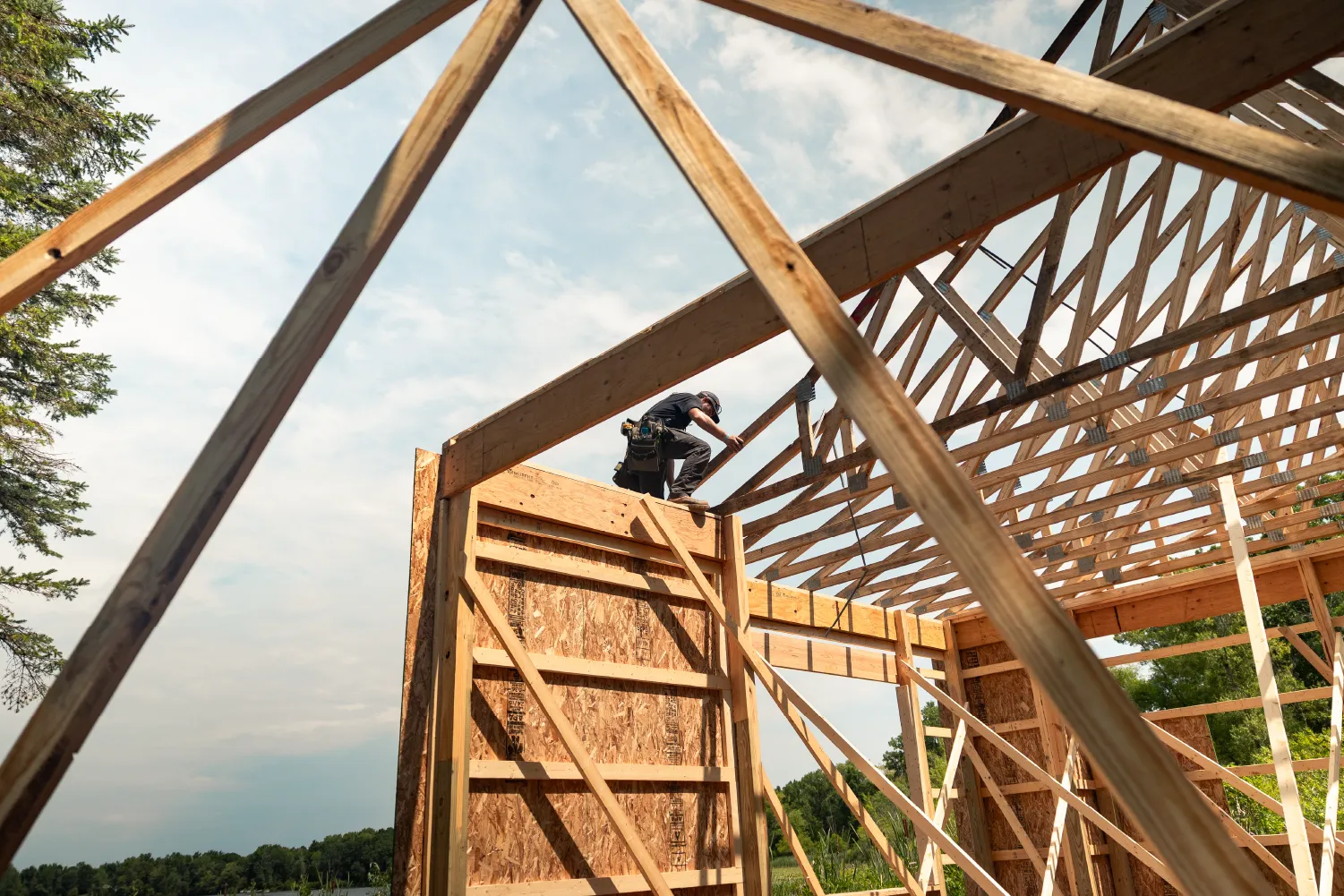 Construction worker positioning wooden beams on the roof frame of a barn under a partly cloudy sky.
