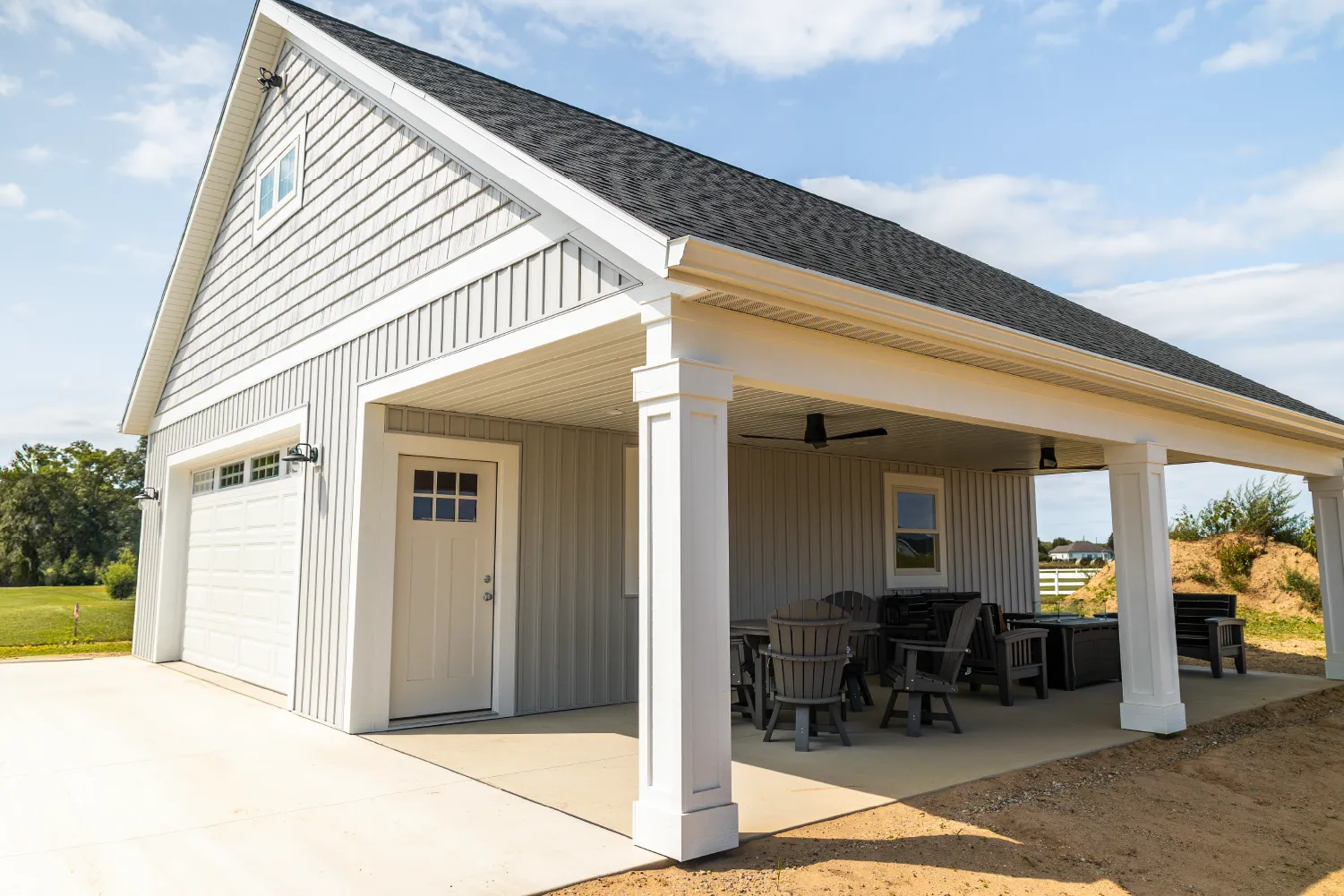 Detached covered patio with outdoor seating and adjacent garage with white door and beige siding under a dark shingled roof.