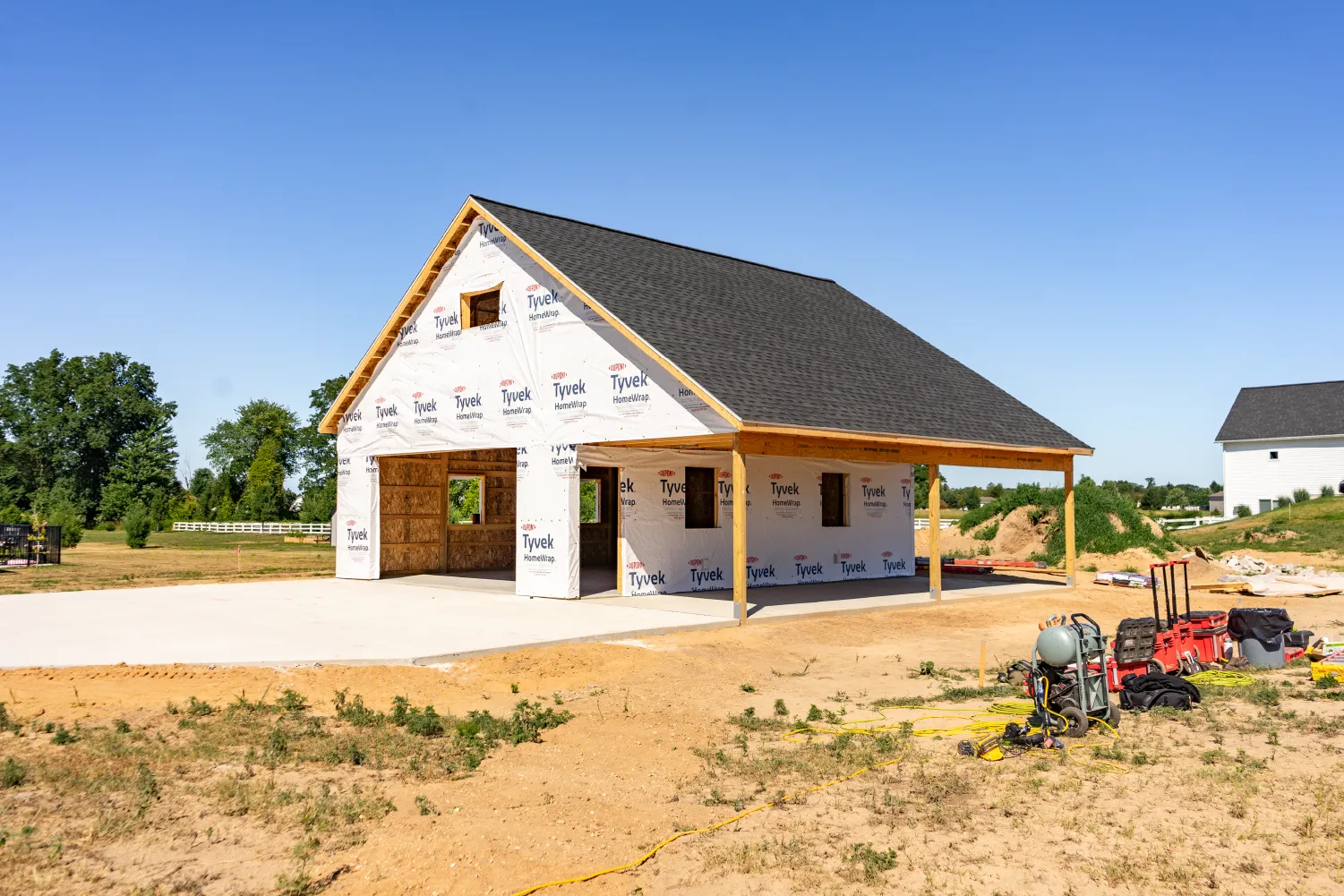 Partially constructed barn covered with Tyvek HomeWrap on a sunny day with construction tools and equipment nearby.