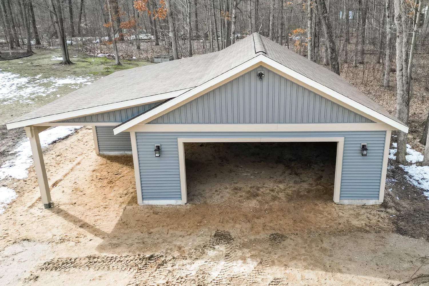 Newly constructed blue two-car garage with an attached open carport in a wooded area with patchy snow on the ground.