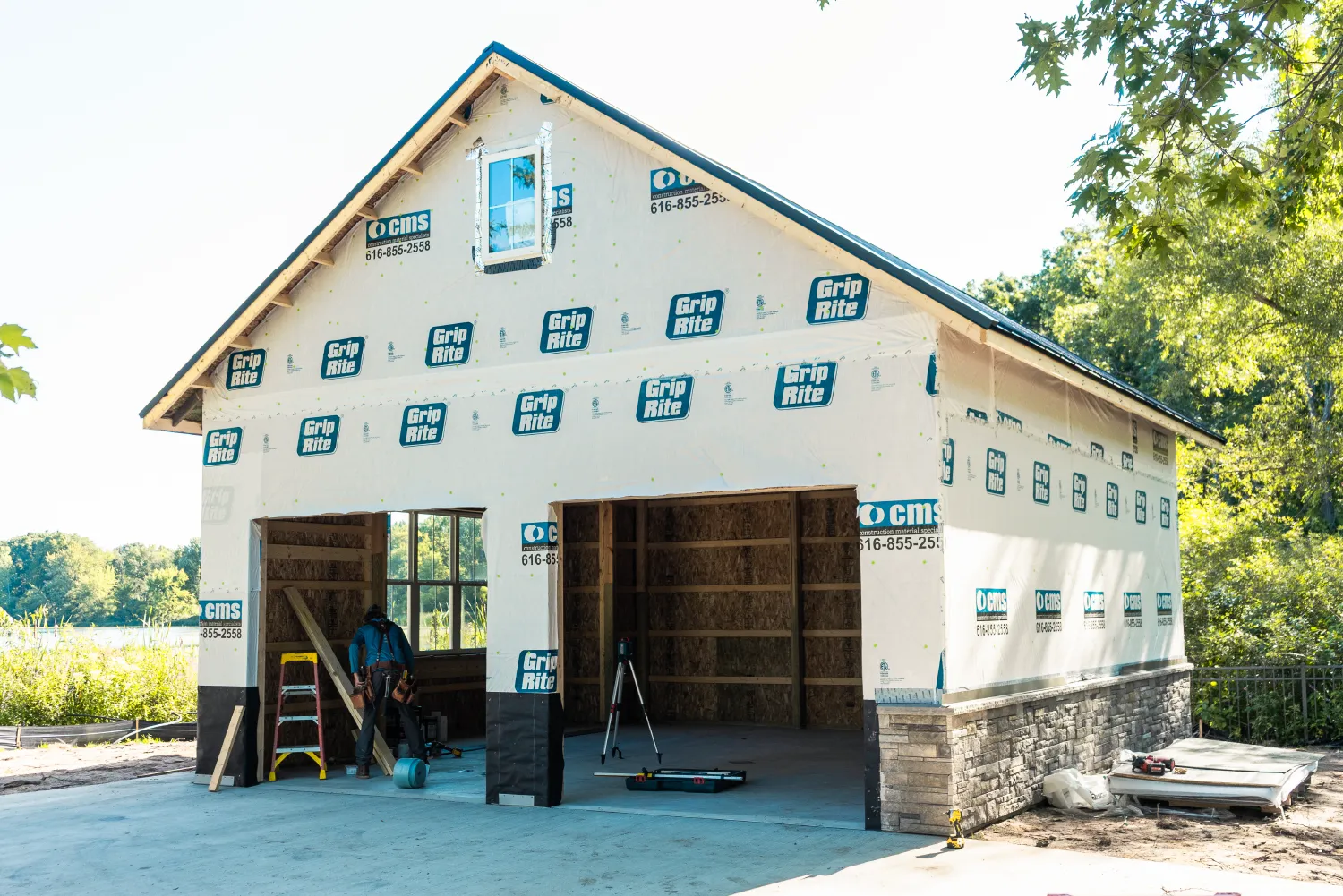 Partially constructed garage with Grip-Rite branded house wrap and a worker inside, surrounded by trees and construction tools.