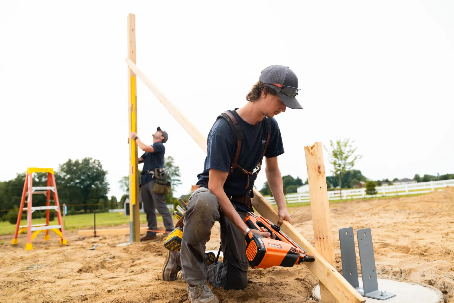 Two construction workers assembling wooden framework outdoors on sandy ground, one kneeling using a nail gun and the other adjusting a vertical wooden beam with a level.