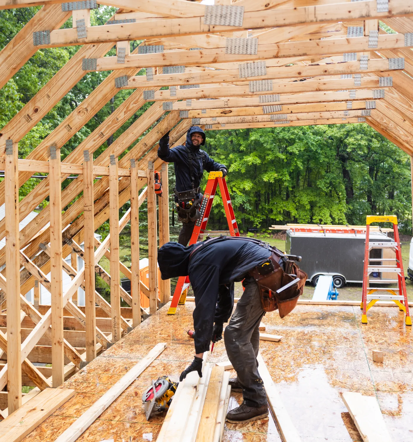 Two construction workers building a wooden frame structure outdoors on a rainy day with trees in the background.