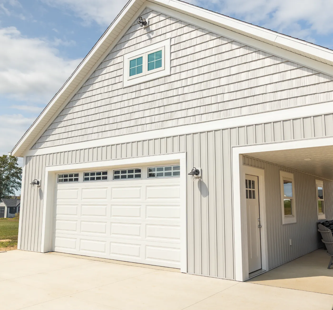 Light gray barn with white trim, a large white garage door with windows, an additional white door, and an adjacent covered patio area.