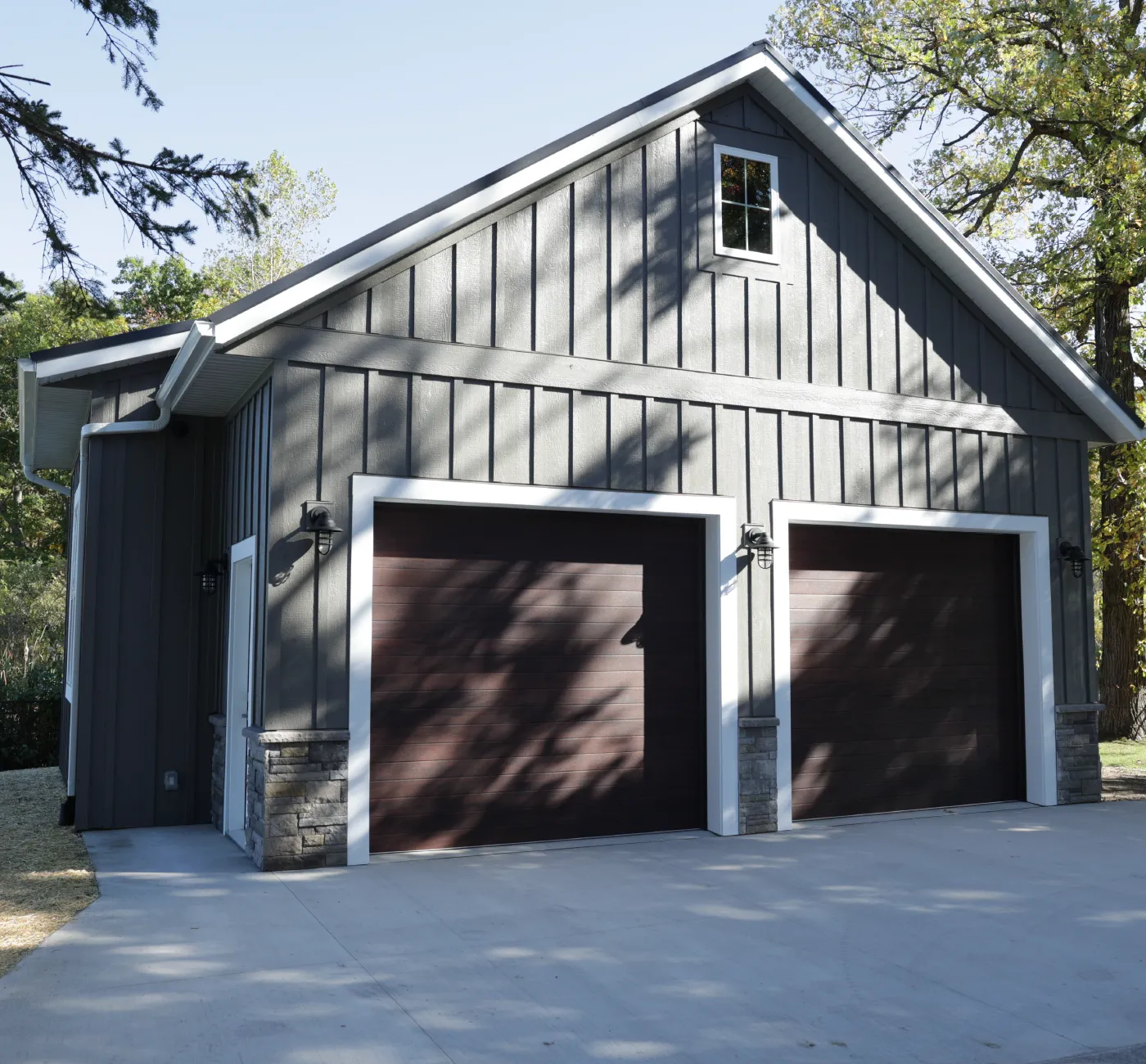 Detached modern barn-style garage with two closed dark brown doors, gray siding, and a small upper window.