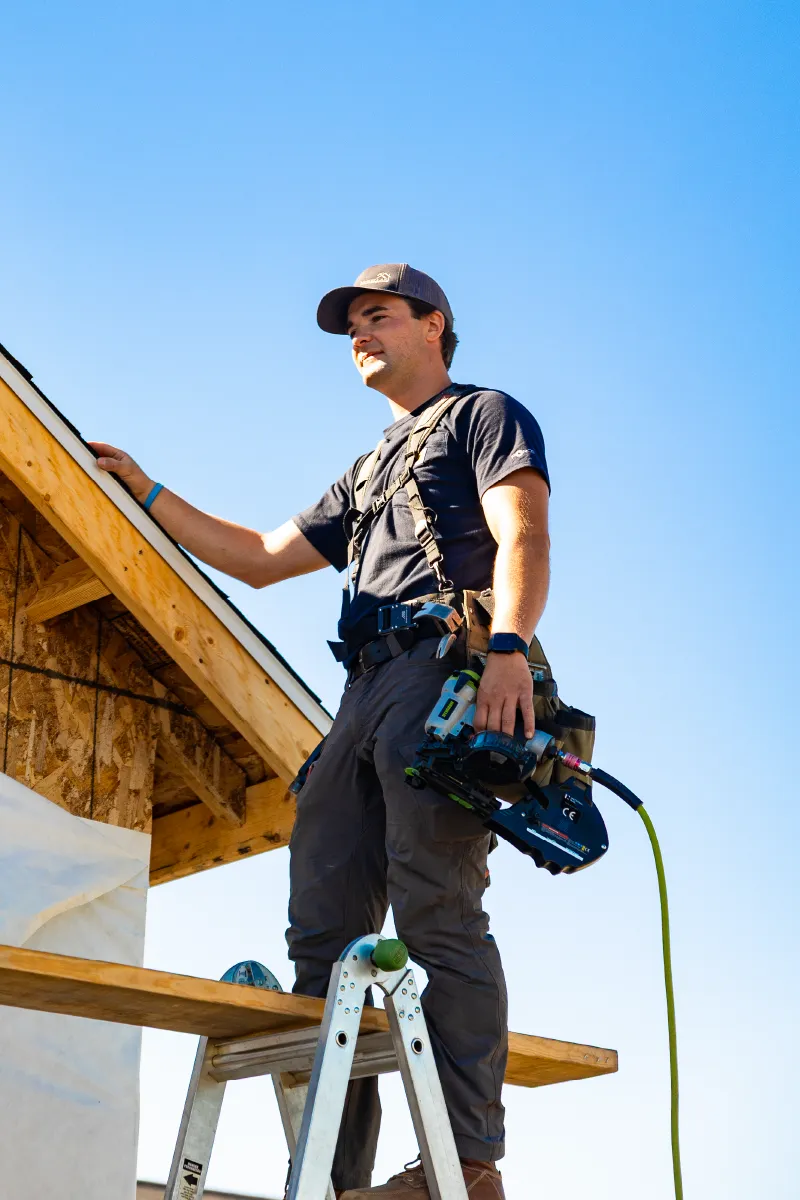 Construction worker standing on a ladder platform holding a nail gun next to a wooden roof frame under a clear blue sky.