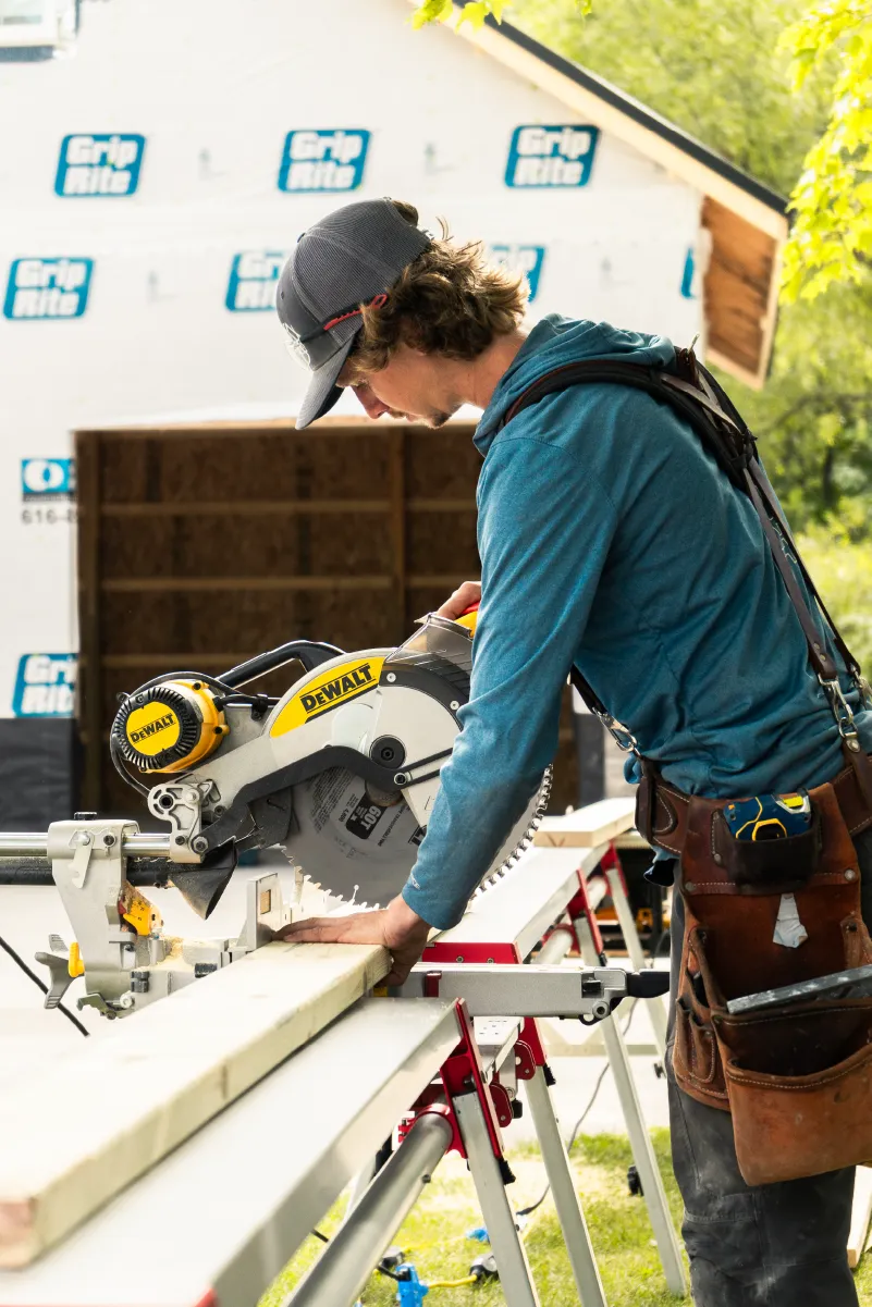 Carpenter wearing a blue hoodie and cap cutting a wooden plank with a DeWalt miter saw outdoors near a building under construction.