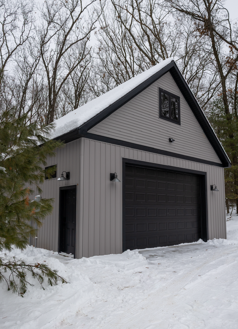 Gray barn with black garage door and snow-covered roof surrounded by leafless trees and snow on the ground.