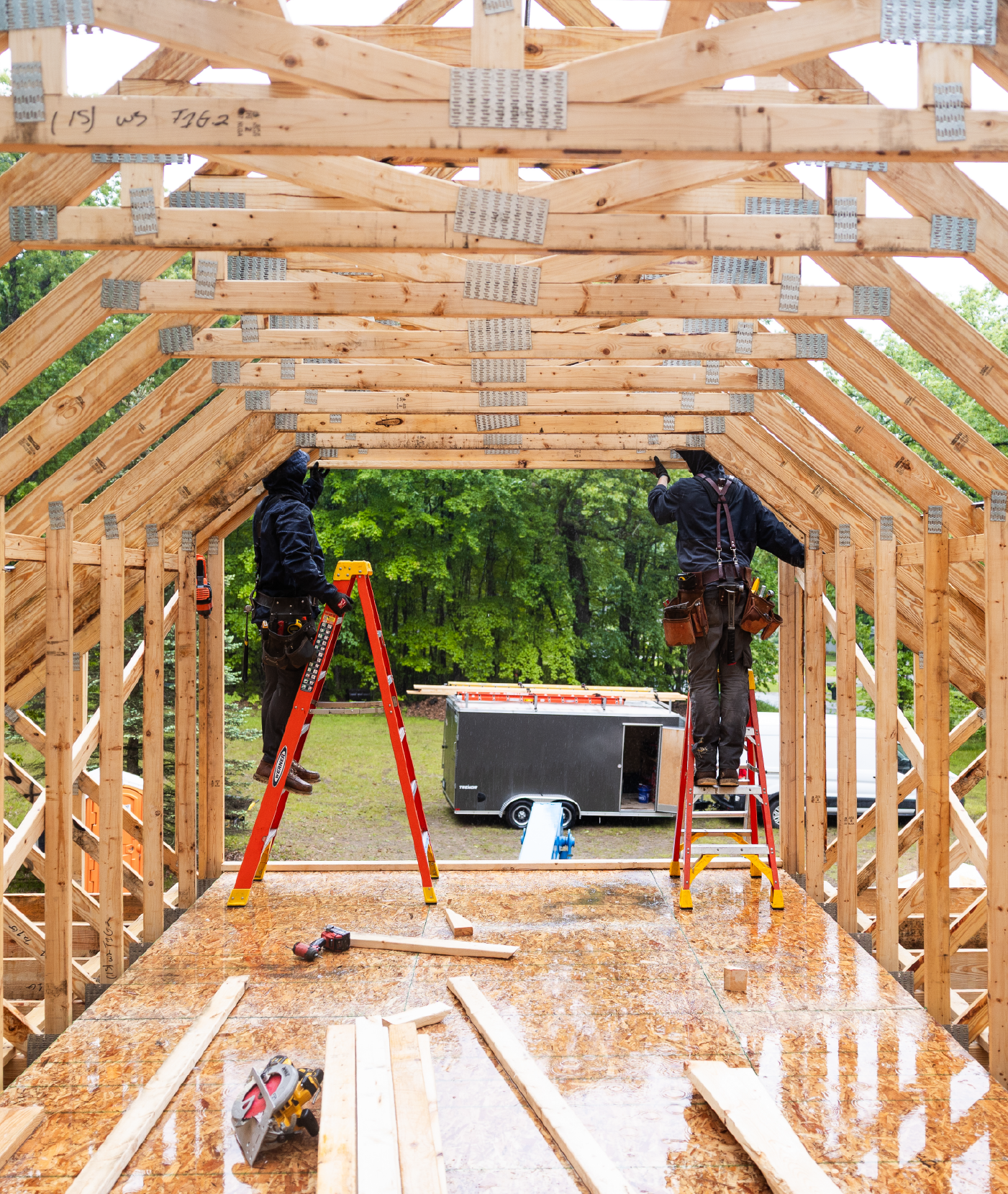 Two construction workers on ladders installing wooden beams inside a partially built wooden structure with green trees in the background.