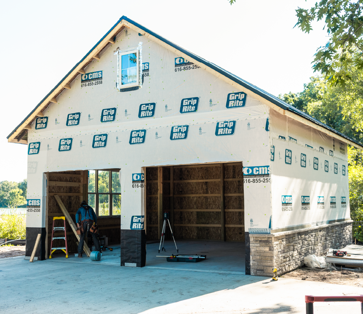 Partially constructed two-car garage with exposed framing and weatherproof wrapping labeled Grip Rite and CMS, a worker is inside on the left side.