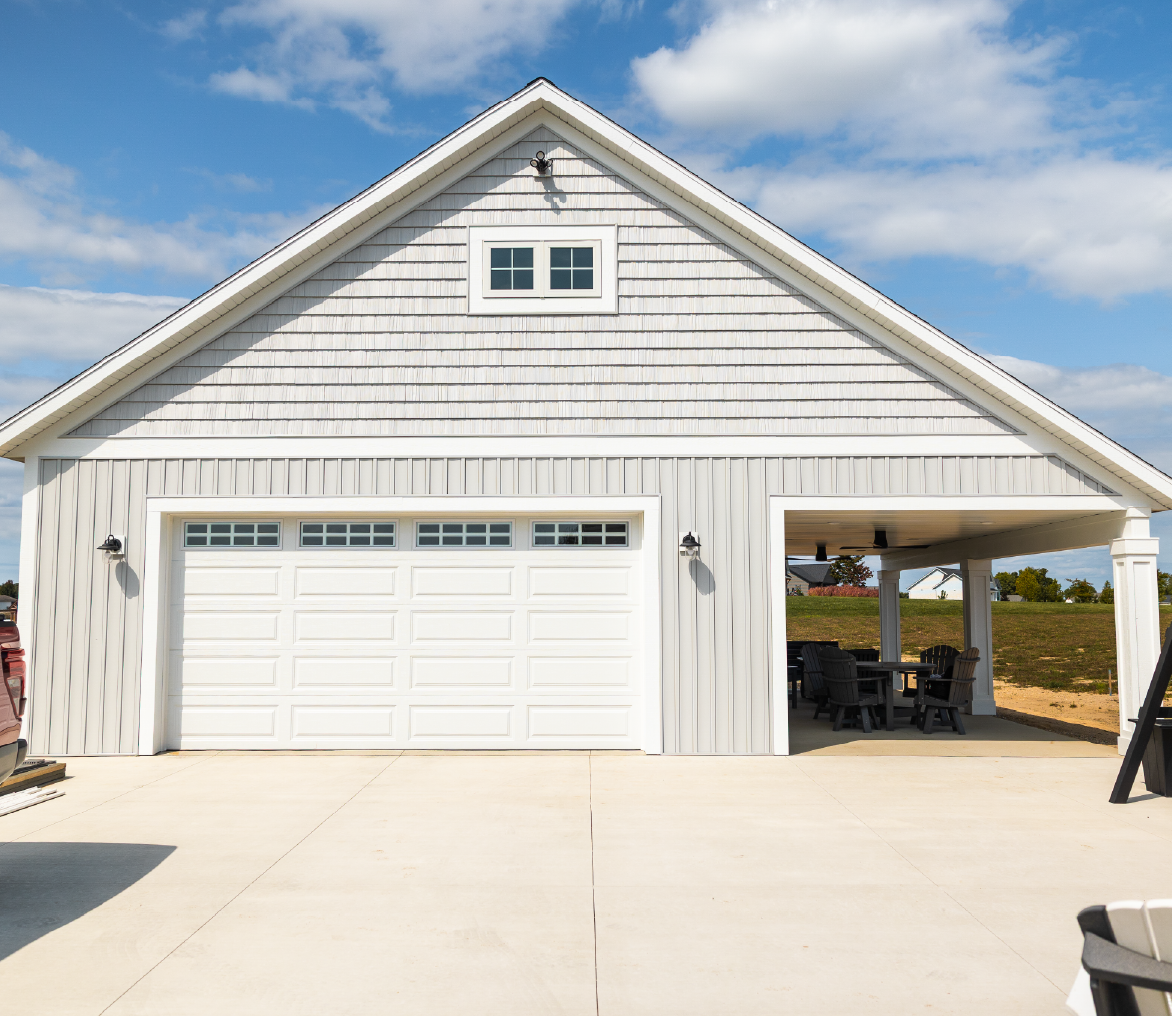 White barn with a closed garage door and an open sided area with outdoor chairs and tables under a blue sky with some clouds.