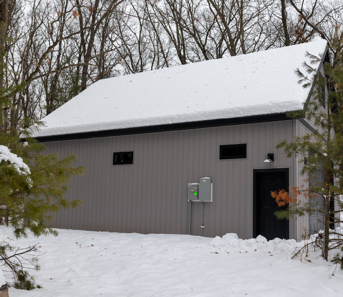 Gray barn with a snow-covered roof surrounded by snow and trees with bare branches.