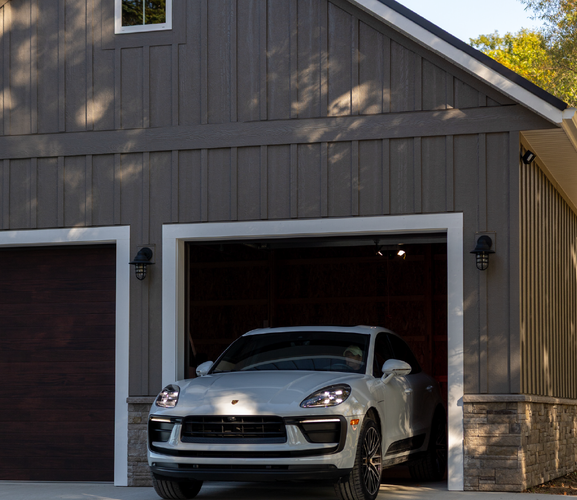 White Porsche car parked halfway out of a two-door garage with gray siding and stone accents during daytime.