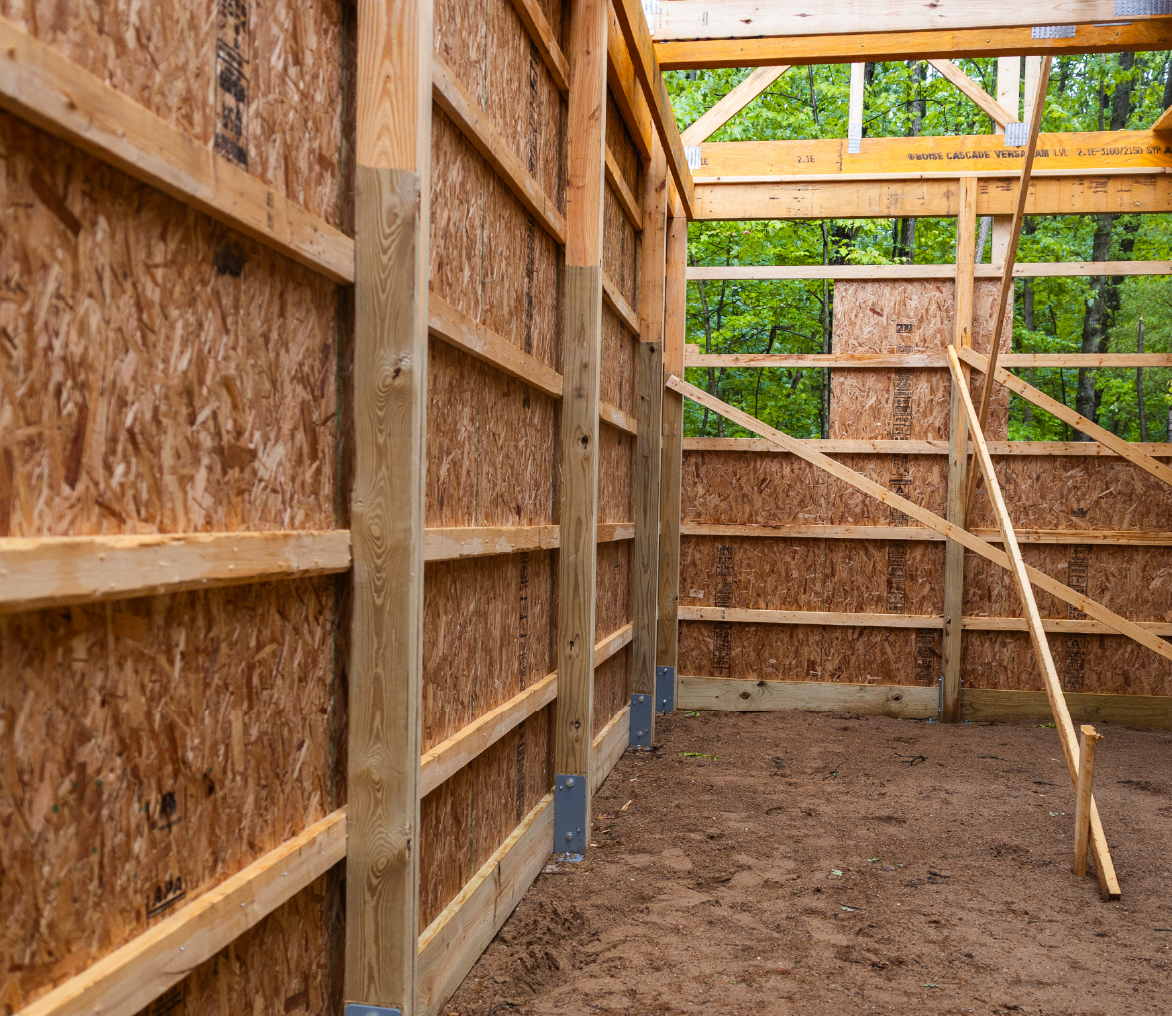 Interior view of a wooden barn under construction surrounded by green trees.