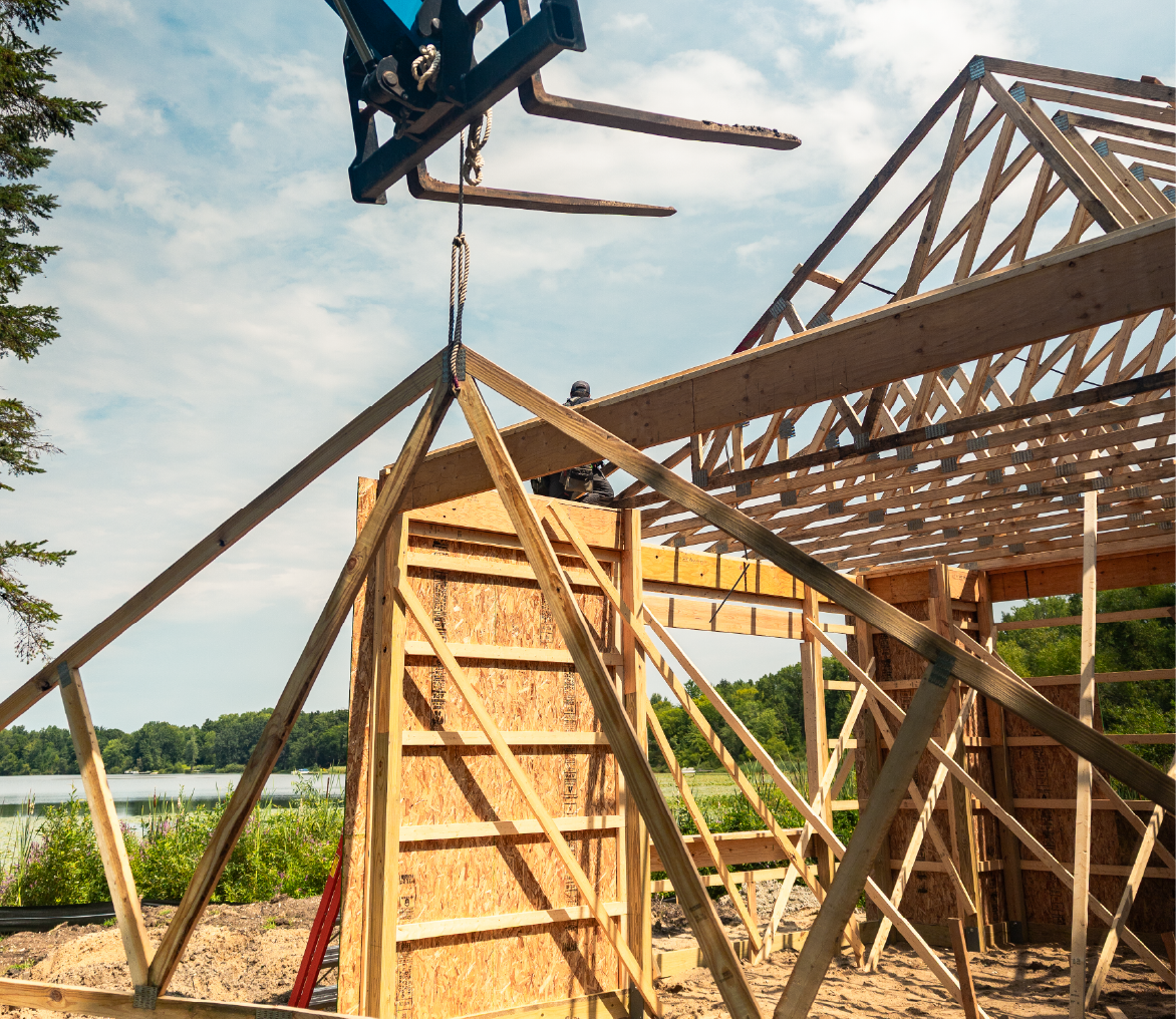Construction site with wooden barn frame being lifted by forklift near a lakeside.