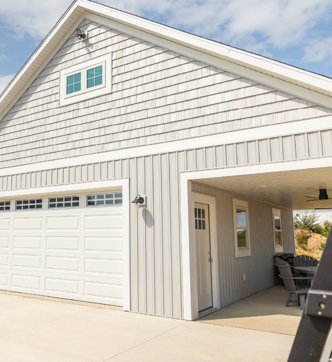 Light gray garage building with a white double garage door, small upper windows, and a covered outdoor seating area with chairs and a table.