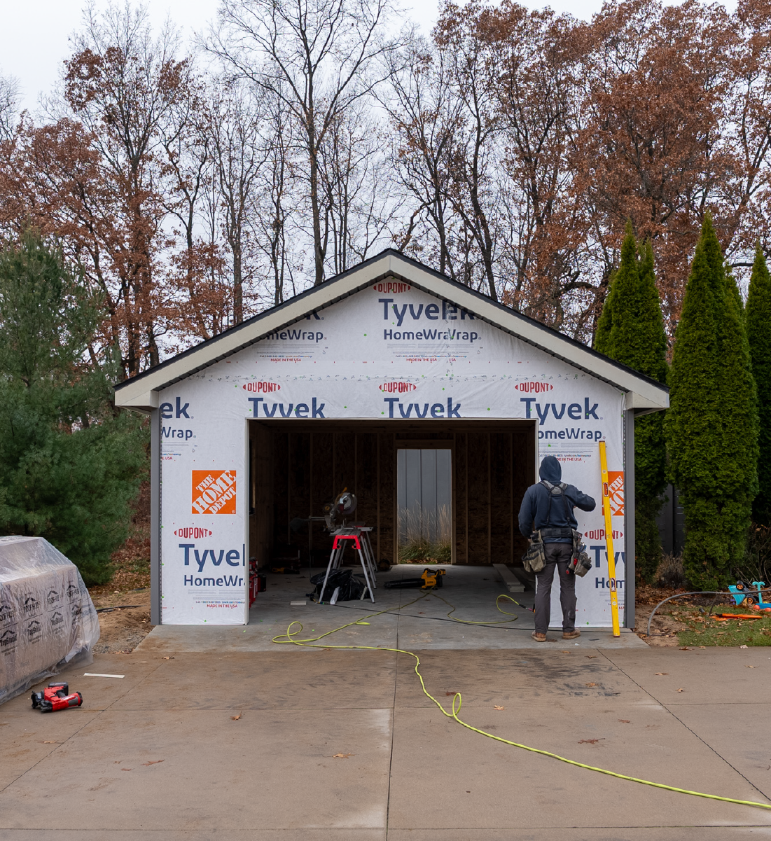 Construction worker standing outside an unfinished garage wrapped in Tyvek HomeWrap with tools and equipment around.