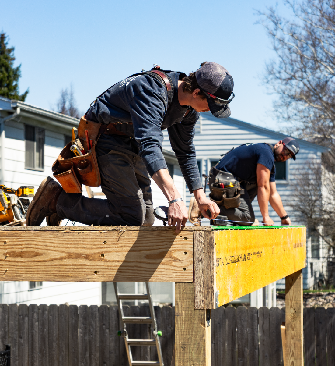 Two construction workers kneeling and measuring on a wooden frame outdoors on a sunny day.