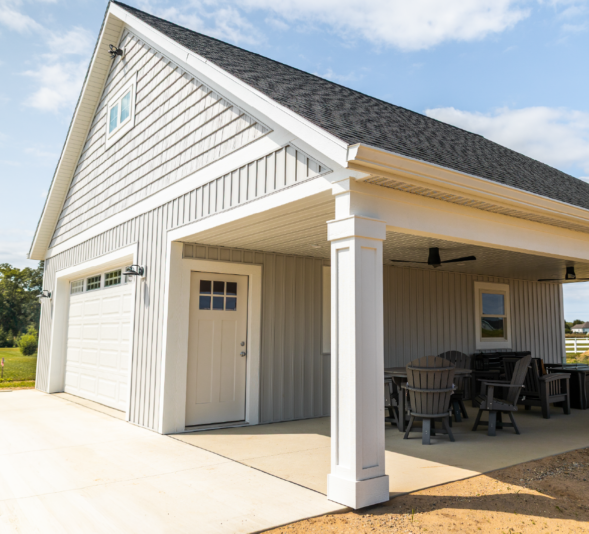 Modern detached garage with white siding, black shingled roof, and covered porch with patio furniture.