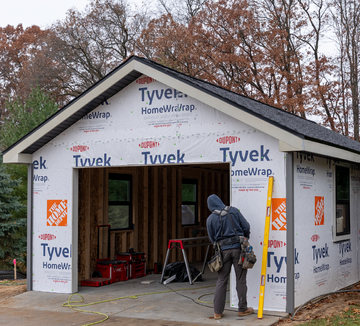 Construction worker inspecting garage framing covered with Tyvek HomeWrap surrounded by autumn trees.