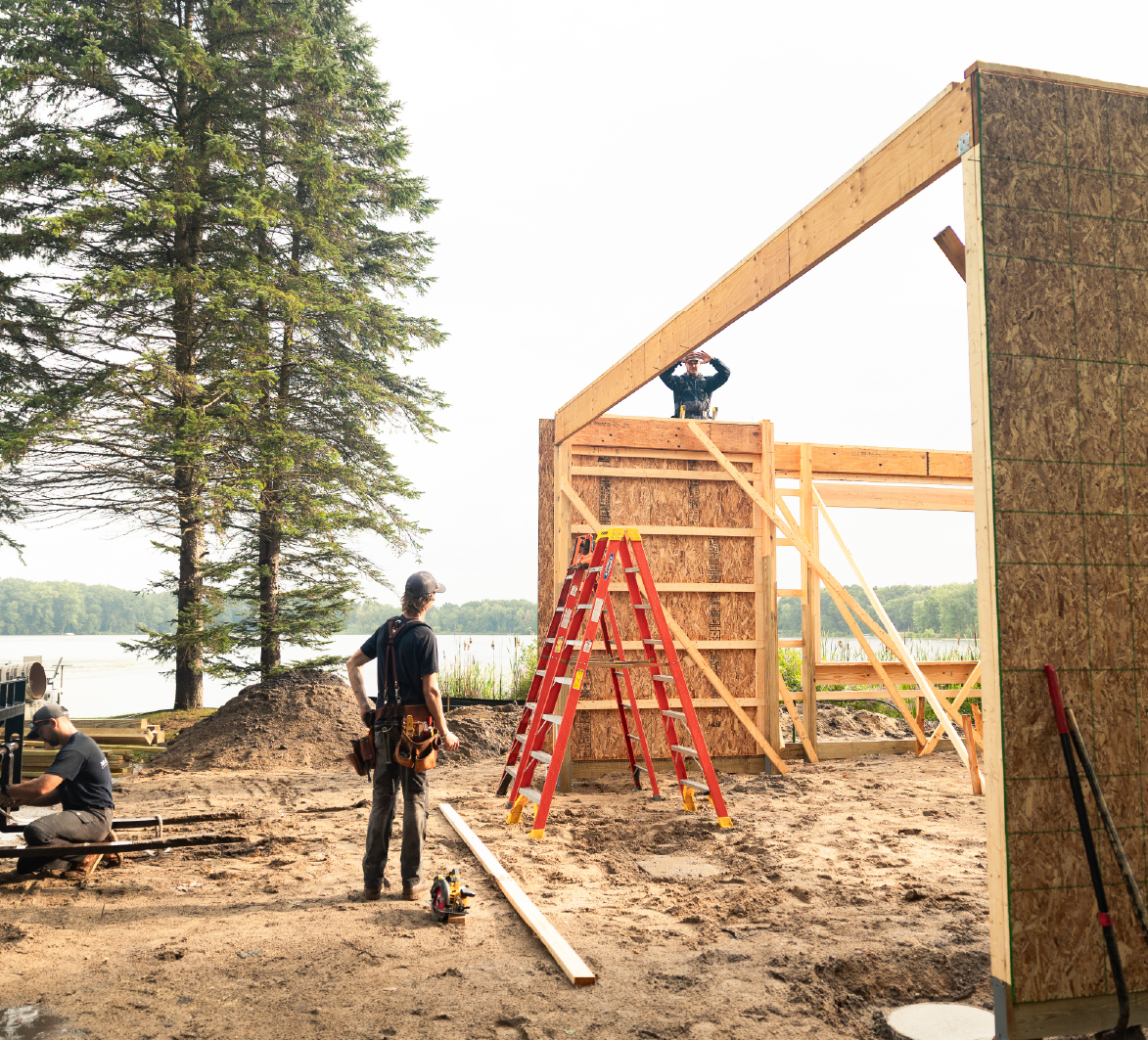 Three construction workers assembling a wooden frame of a building near a lakeside with tall trees in the background.