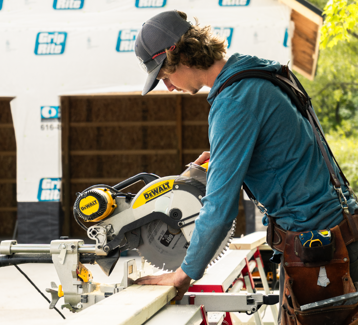 Construction worker in blue hoodie using DeWalt miter saw to cut wooden plank at outdoor building site.