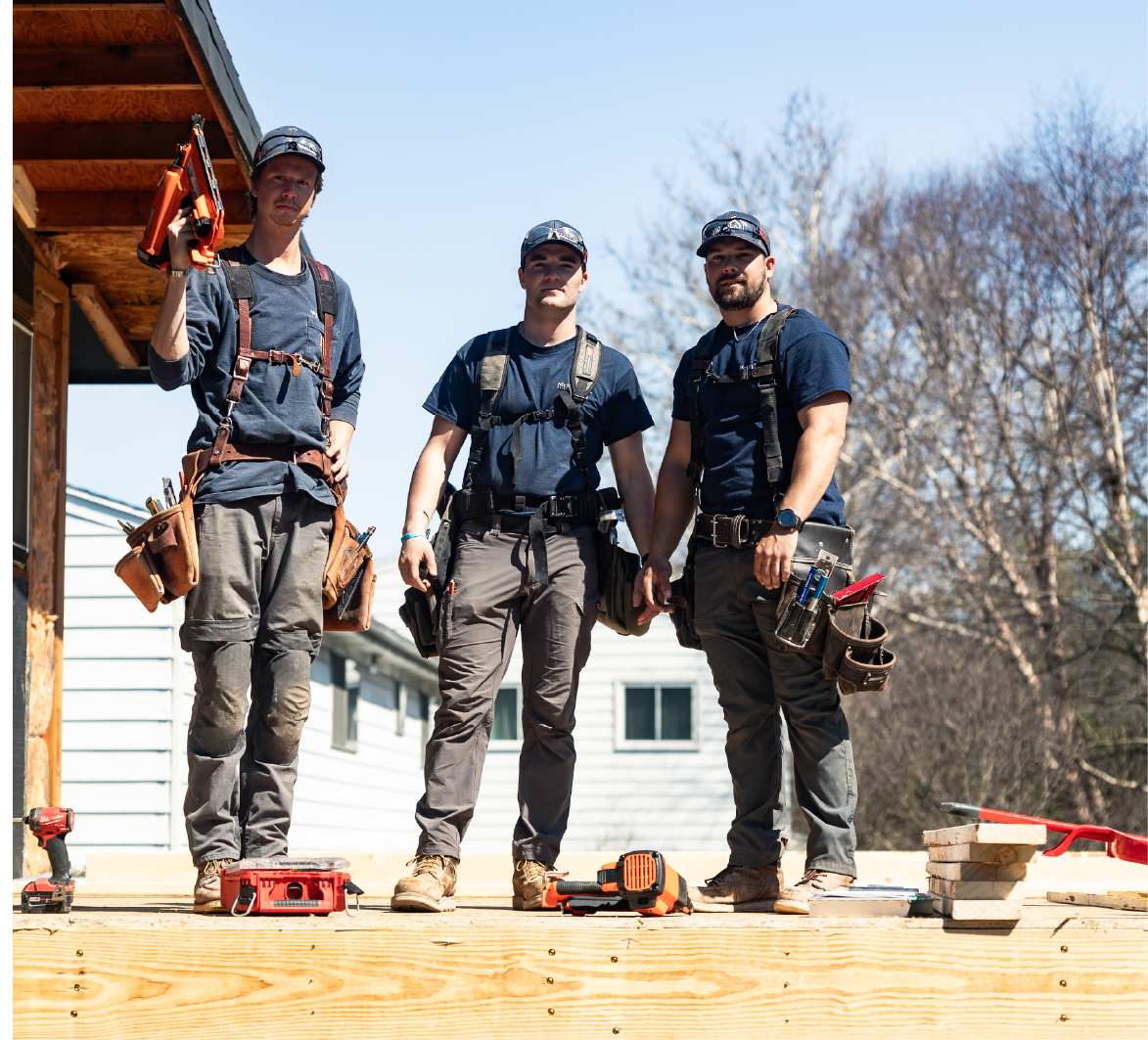 Three construction workers standing on a wooden platform outdoors with tools and equipment, wearing tool belts and caps.