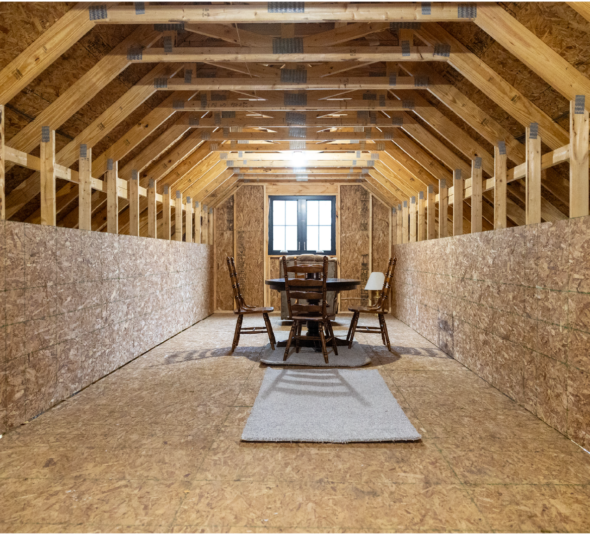 Interior of an unfinished barn with wood framing and oriented strand board walls, featuring a round table and four wooden chairs near a window.