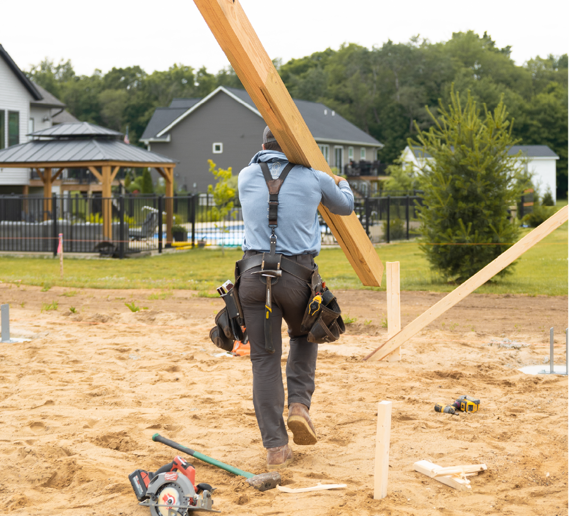 Construction worker in a harness carrying a wooden beam on a sandy site with houses and greenery in the background.