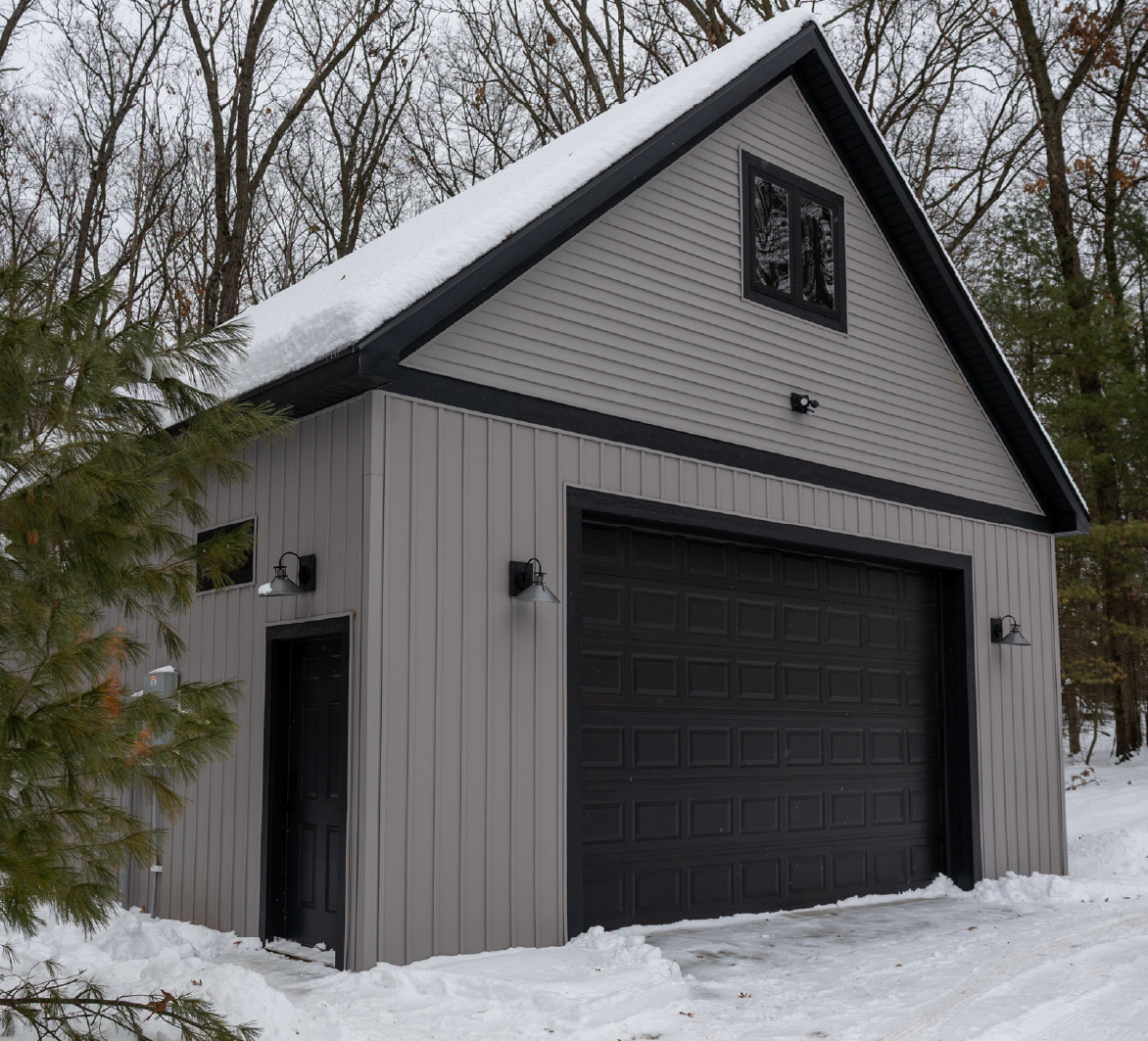 Gray barn-style building with black garage door and side door, snow-covered roof, and surrounding snow and trees.