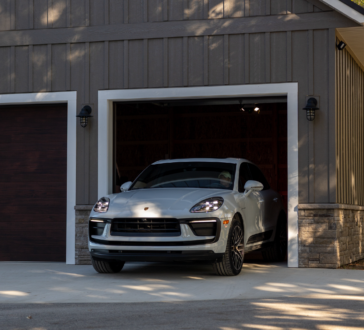White Porsche Macan SUV partially parked inside a garage with wooden exterior walls and stone accents.