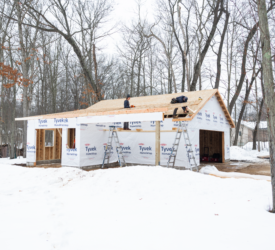 Two workers building the wooden roof frame of a house under construction surrounded by snow and leafless trees.