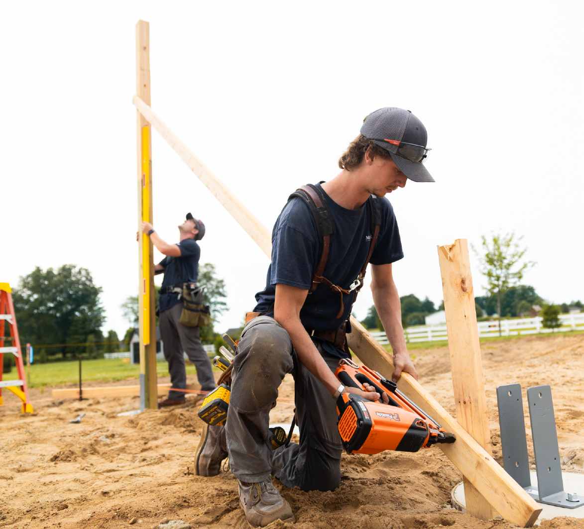 Construction workers installing wooden beams outdoors on a sandy ground with tools and equipment.