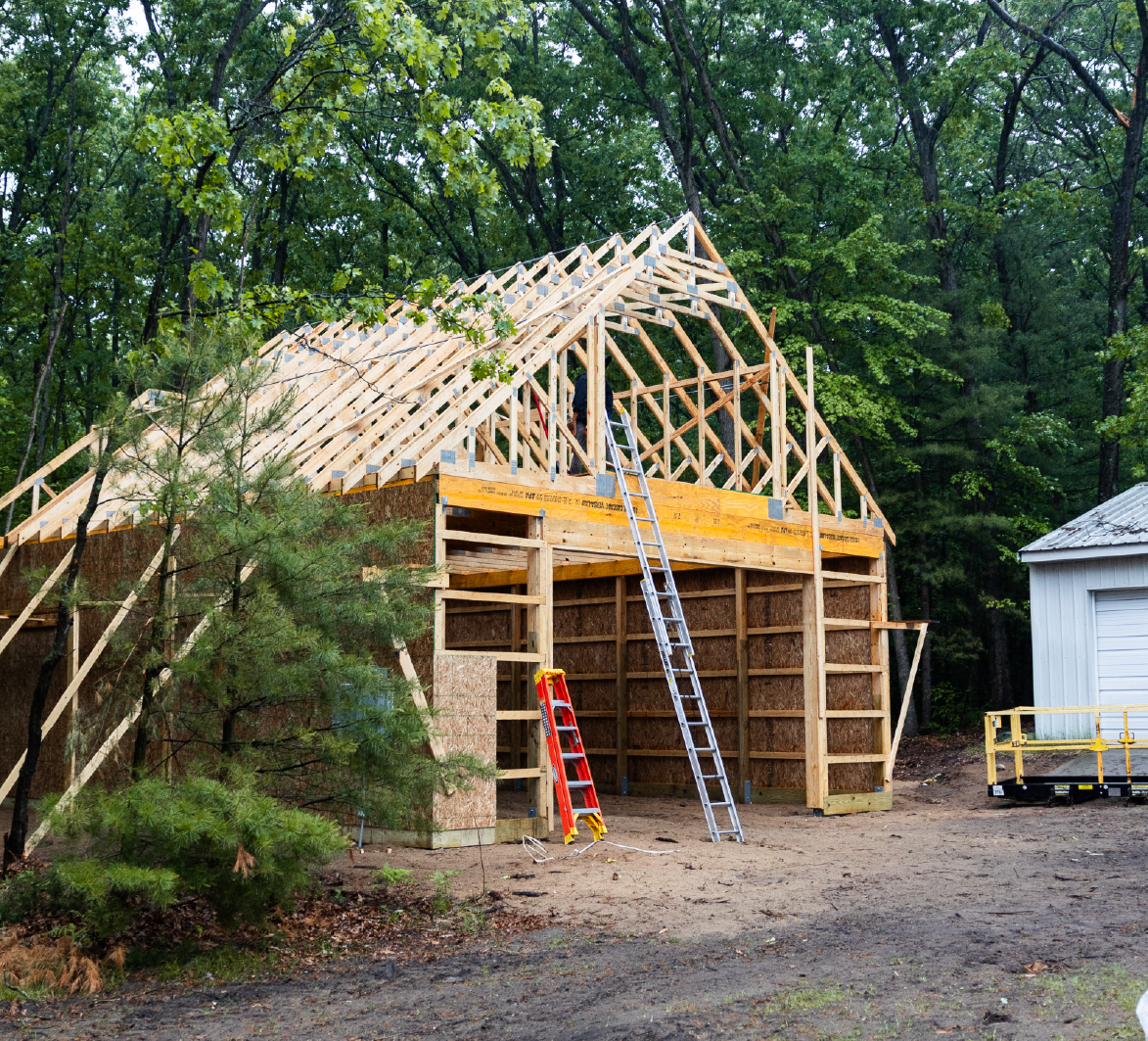 Wooden barn structure under construction surrounded by green trees with ladders placed against it.