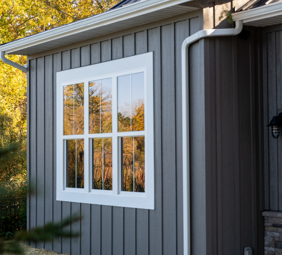 White-framed window with reflections of autumn trees on the gray vertical paneled exterior wall of a house.
