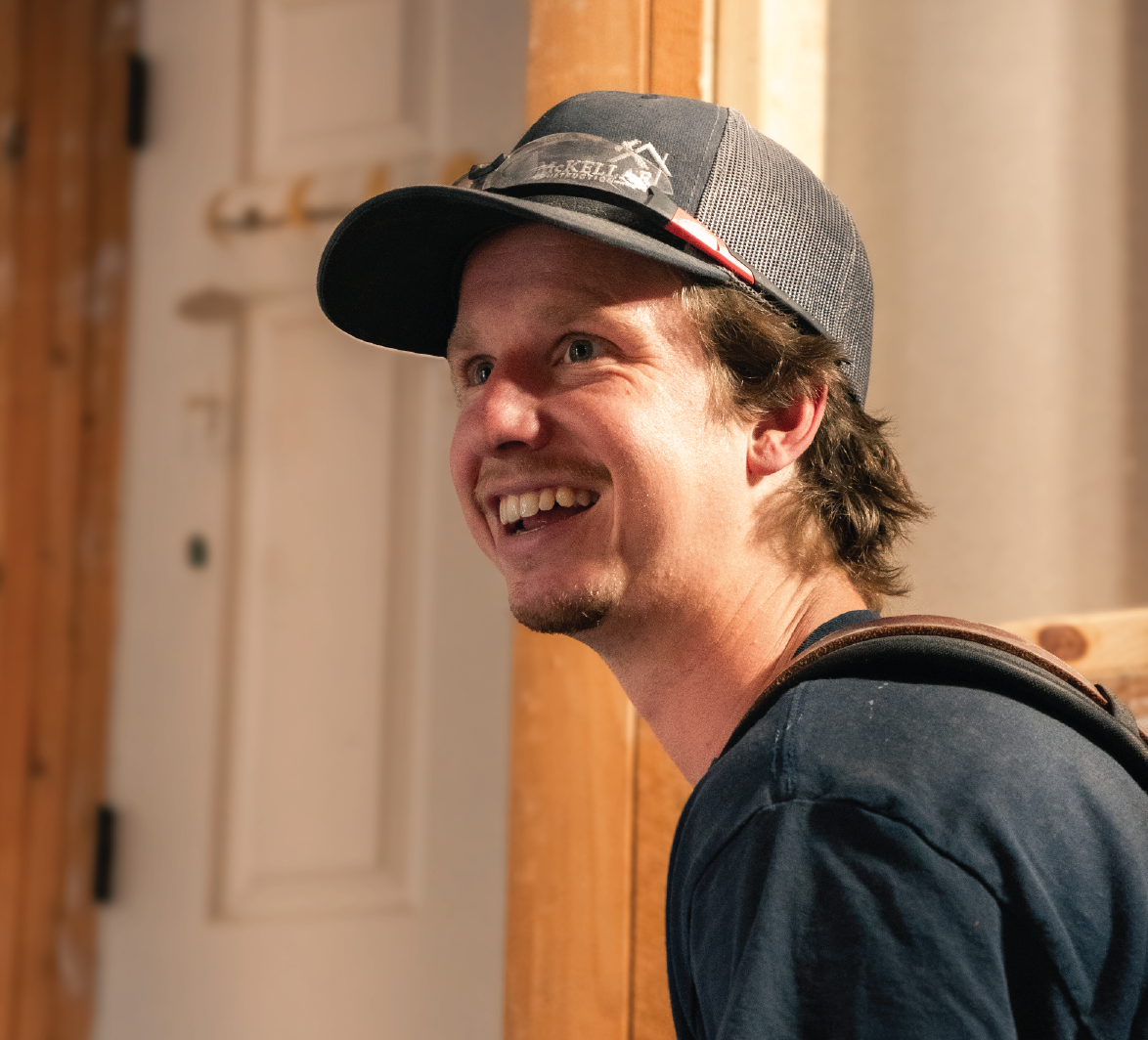 Smiling man wearing a black cap and dark shirt indoors with wooden walls in the background.
