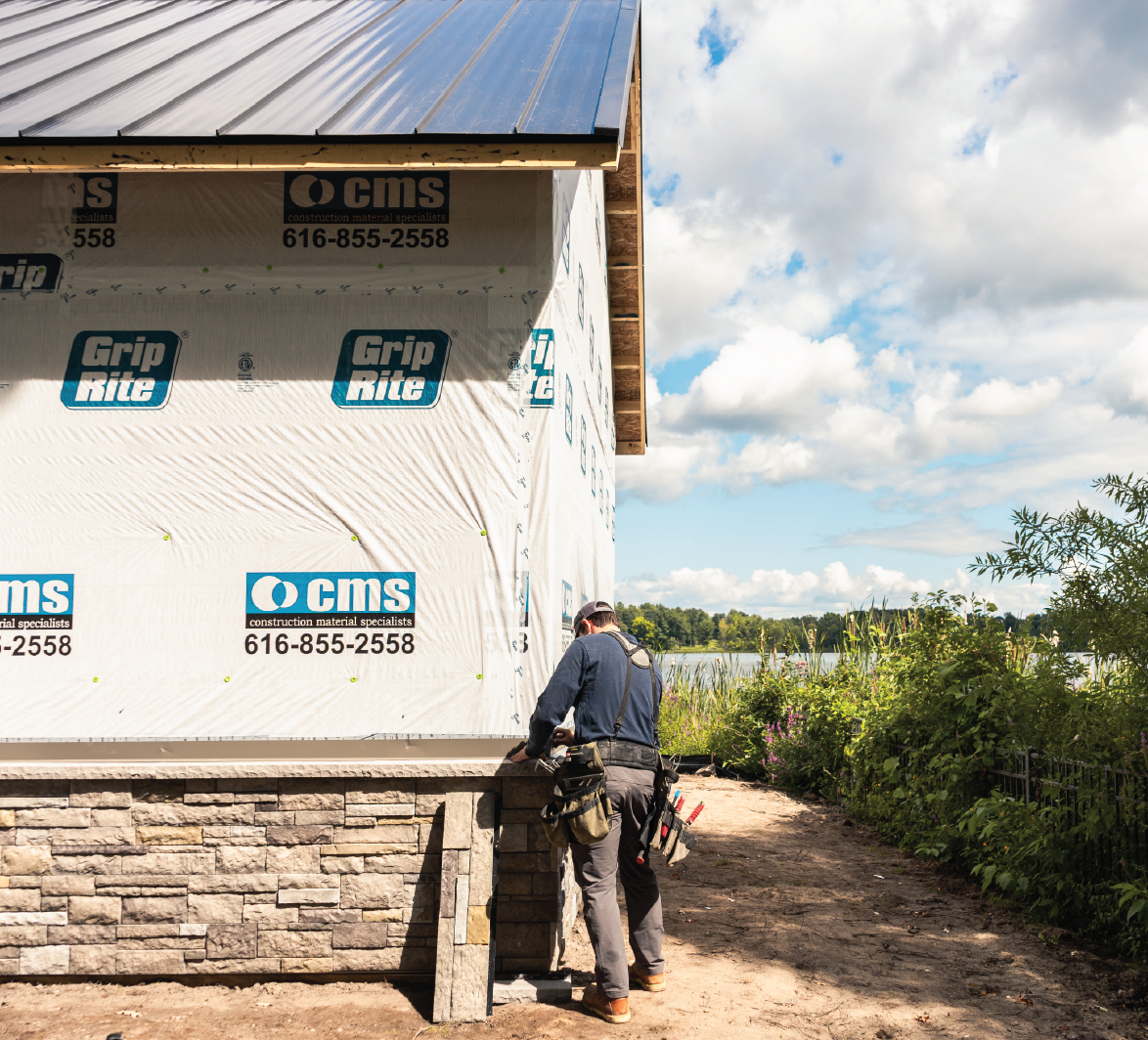 Construction worker installing stone veneer on the exterior wall of a building under construction near a lake and greenery.