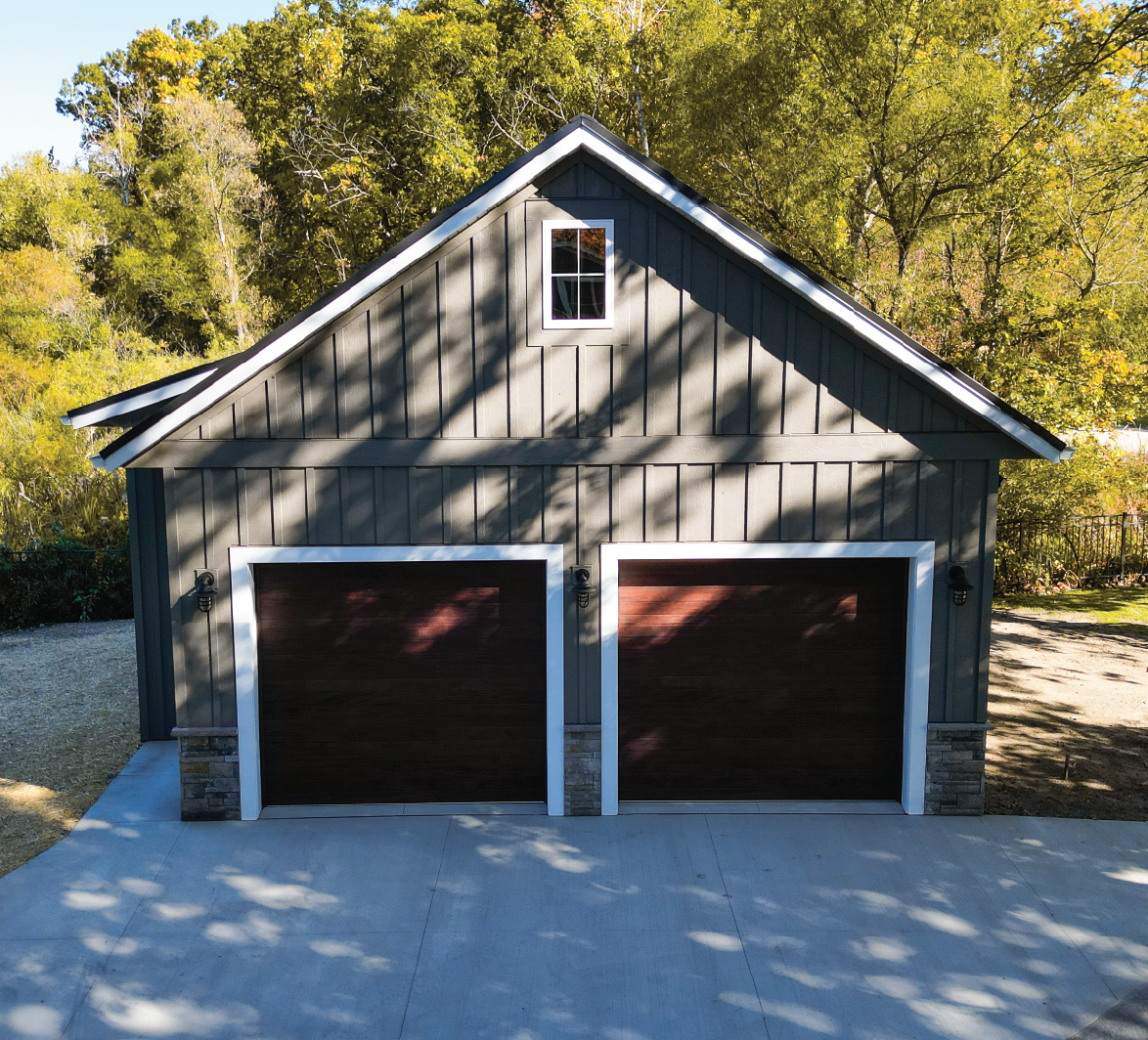 Gray two-car garage with dark wooden doors, white trim, and a small window above, surrounded by green trees.