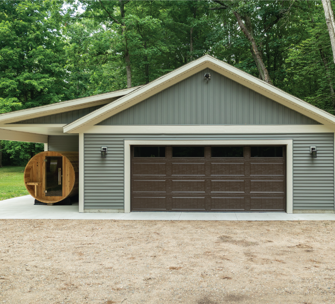 Gray garage with dark brown door attached to a forest surrounded house, with a wooden barrel sauna on the left side.