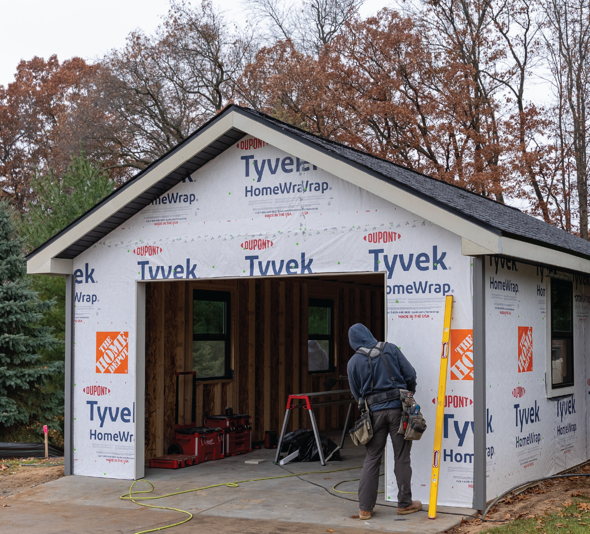 Construction worker inspecting a partially built garage wrapped with Tyvek HomeWrap on a concrete slab driveway.