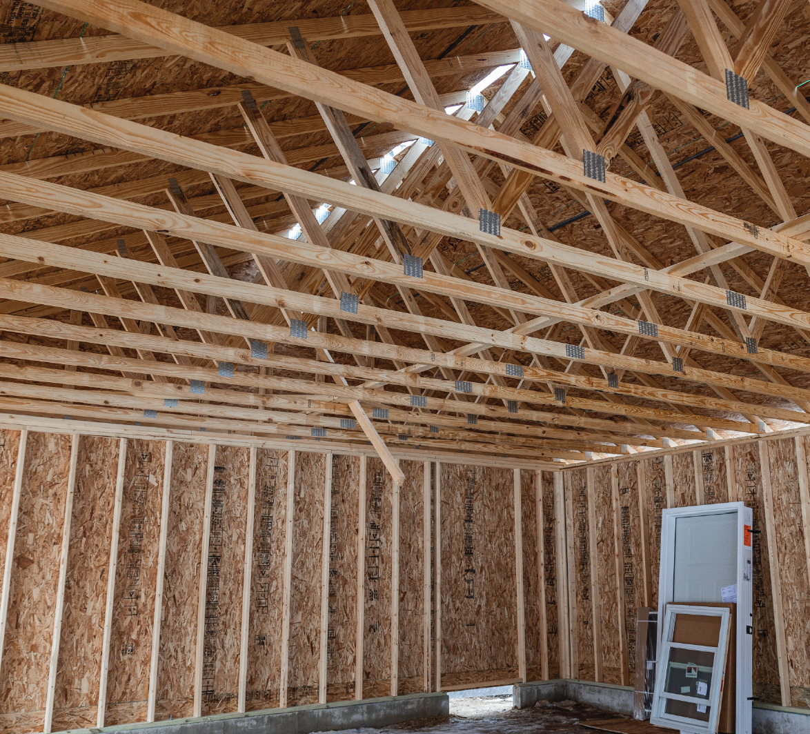Interior view of a wooden building frame under construction, showing roof trusses and wall studs with construction materials stacked inside.