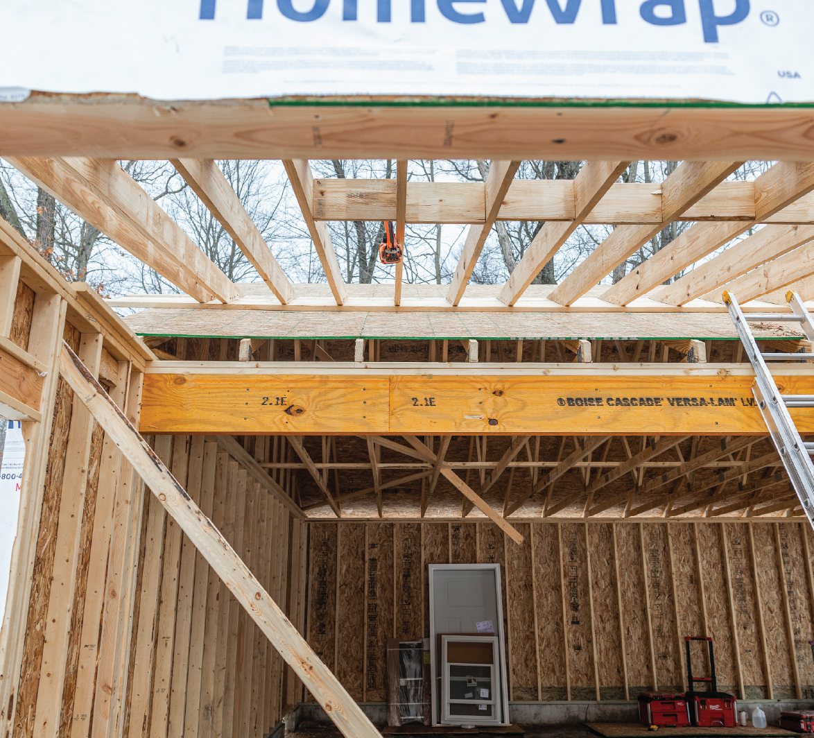 Interior view of a wooden house frame under construction showing exposed beams, plywood sheathing, and construction tools.
