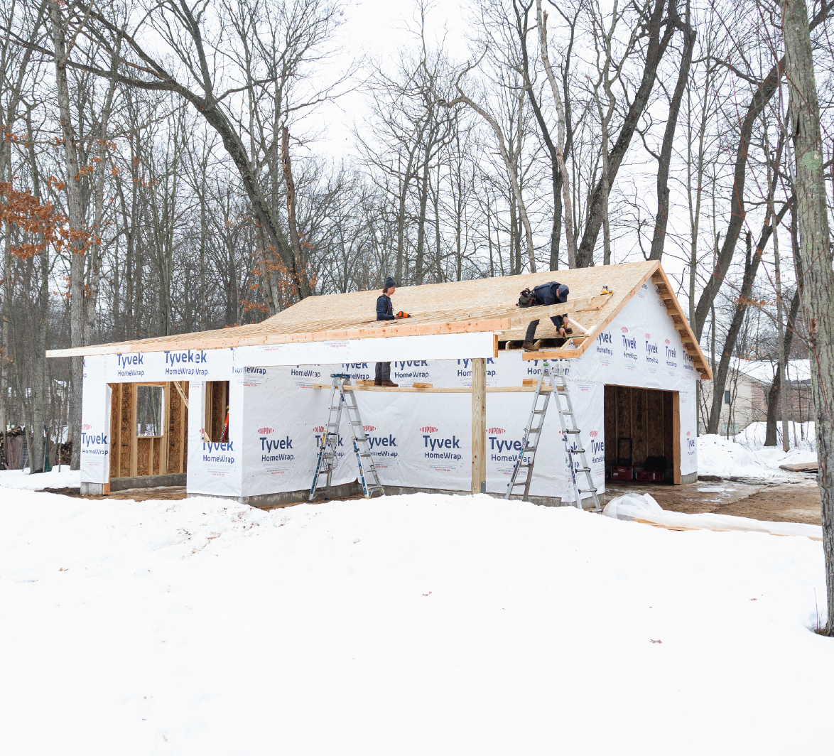 Two construction workers building a wooden roof frame on a snow-covered site surrounded by leafless trees.