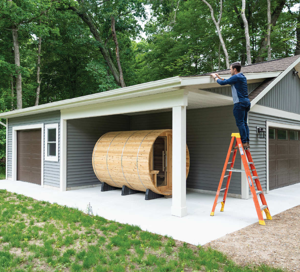Man standing on an orange ladder working on the roof overhang of a gray house with two brown garage doors and a wooden barrel sauna under the carport.